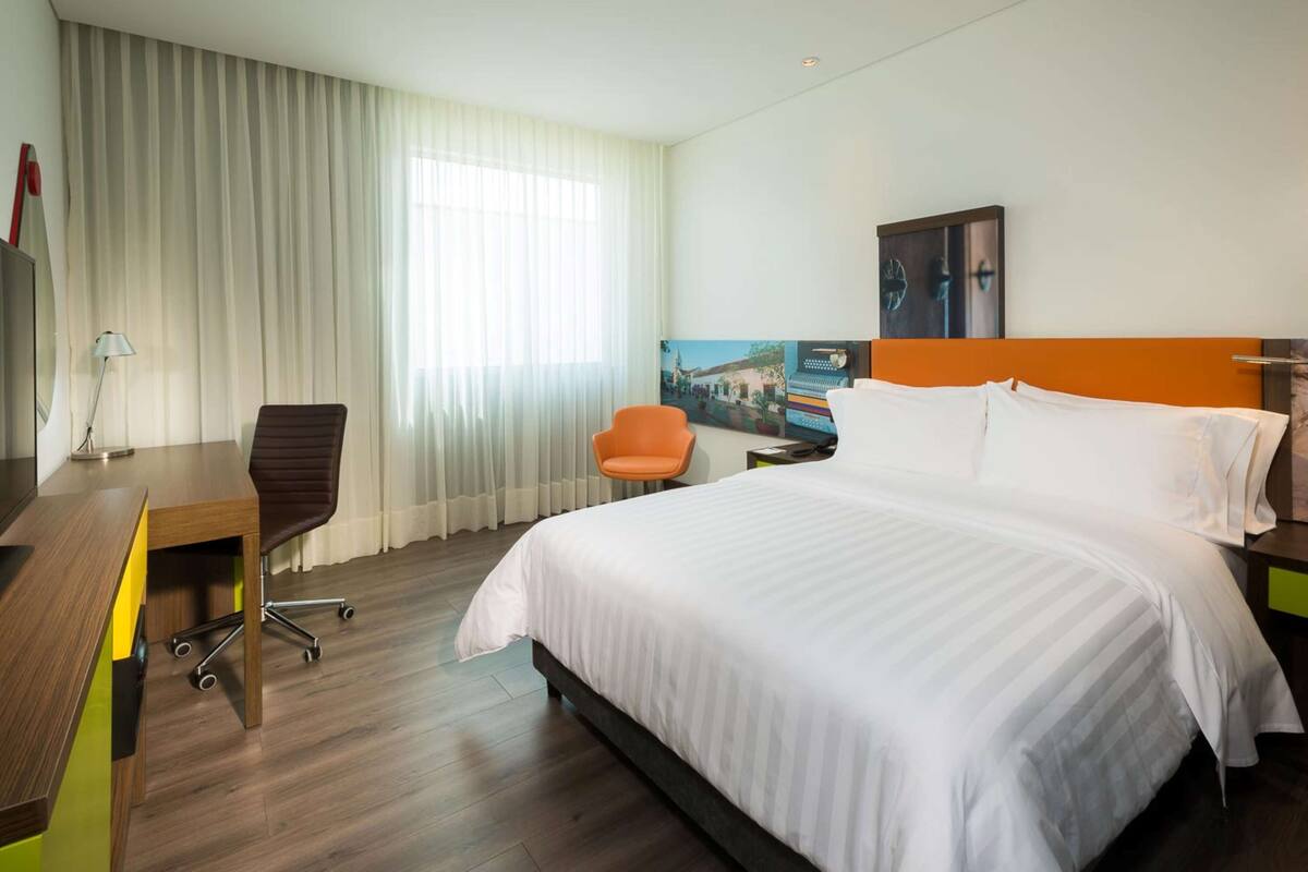 A hotel room with a bed, a desk, an orange chair, and a large window with curtains.