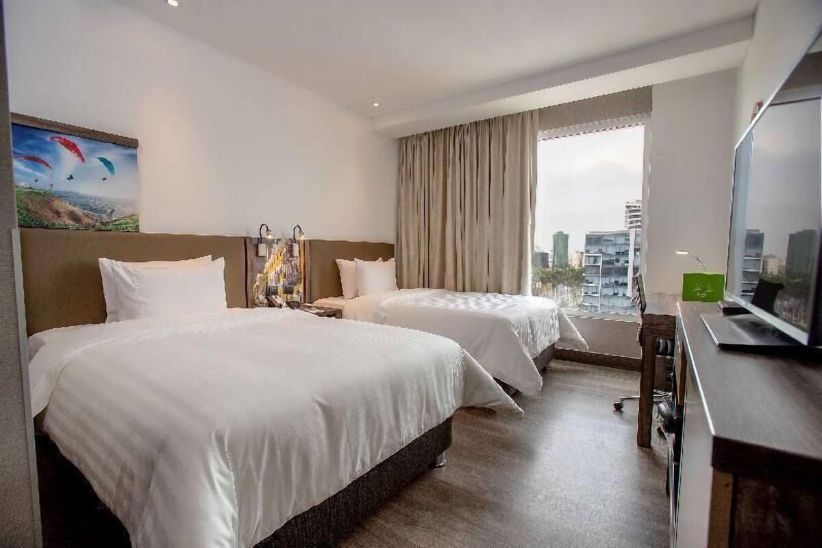Habitación de hotel con dos camas, un escritorio con computadora, televisor y una ventana grande con vista a la ciudad.