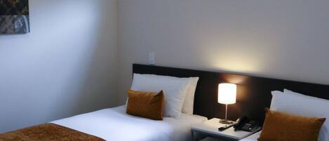 A hotel room with two beds, each with white linens and brown throw blankets. There is a nightstand with a lamp and a telephone between the beds. A window above the beds allows natural light into the room.