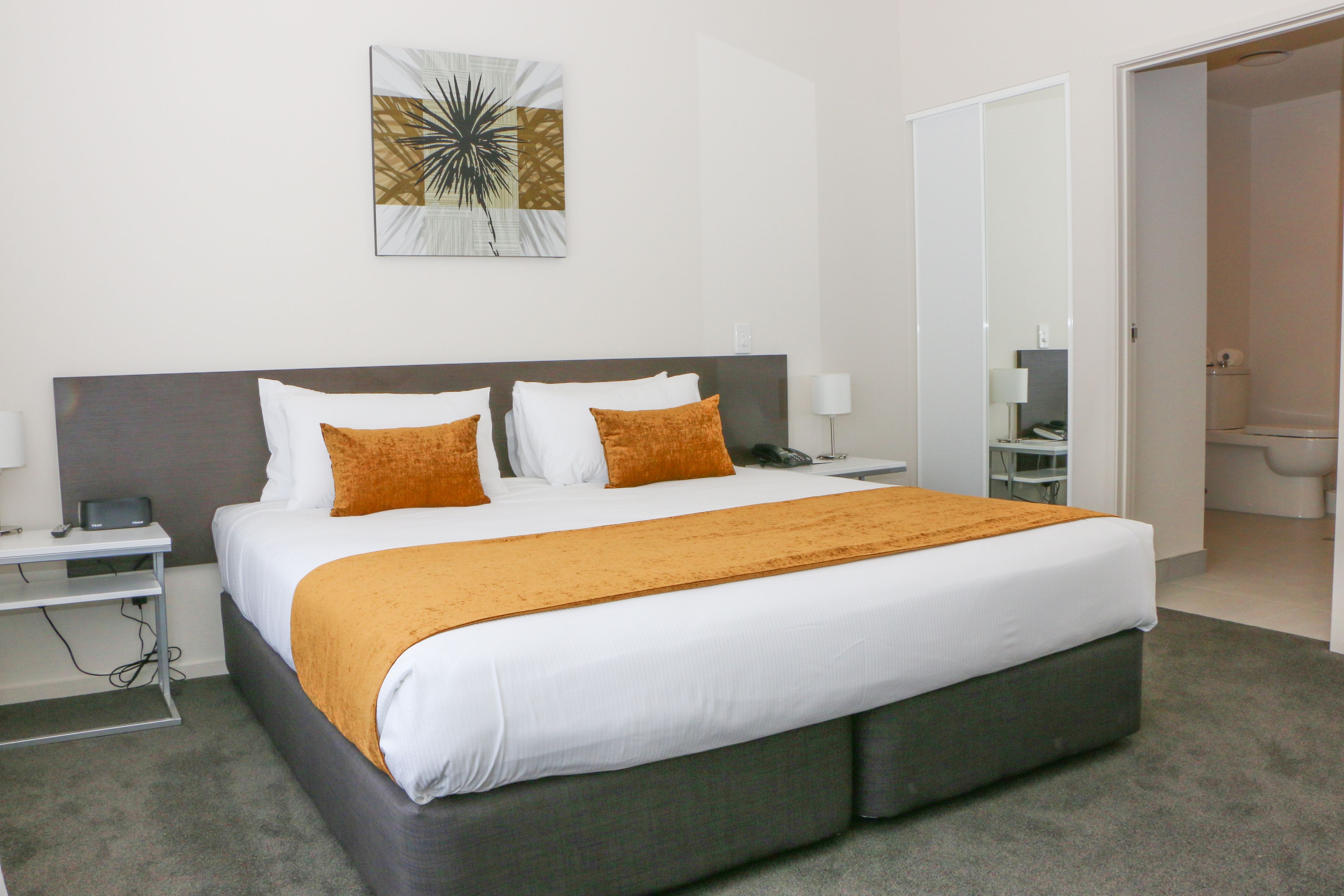 Quarto de hotel moderno com uma cama grande, uma mesinha de cabeceira, uma luminária e uma obra de arte na parede, além de um banheiro visível por uma porta aberta.