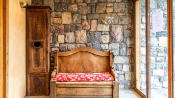 Una panca di legno con un cuscino rosso decorato, addossata a un muro in pietra, una porta in legno e una finestra con cornice in pietra.