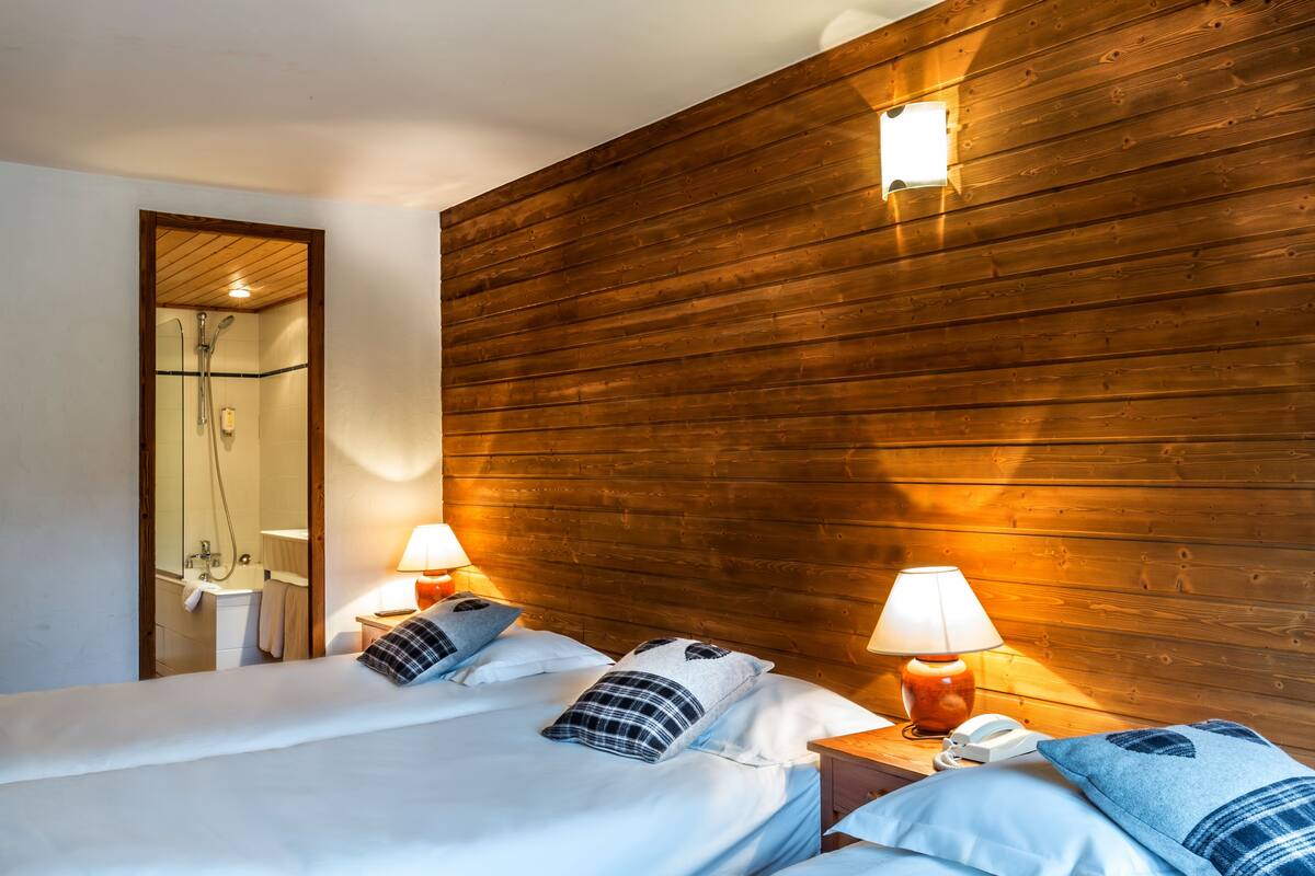 A hotel room with wooden paneling, two beds with white linens, two bedside lamps, a mirror, and a shower area.
