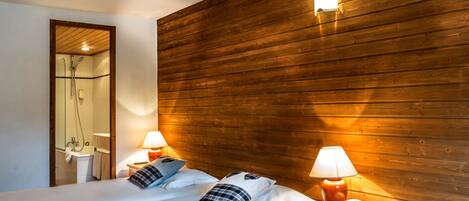 A hotel room with wooden paneling, two beds with white linens, two bedside lamps, a mirror, and a shower area.