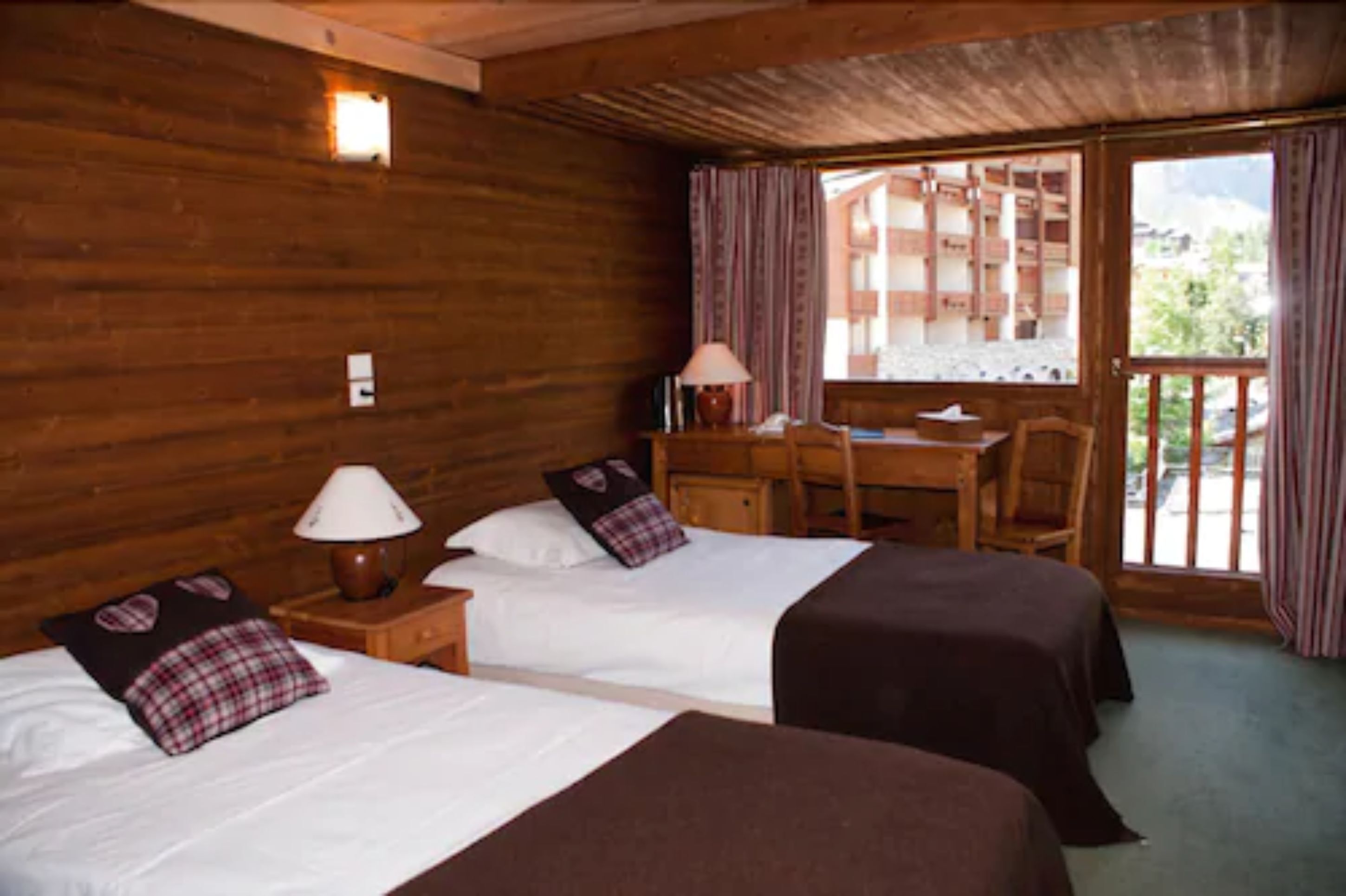 A wooden cabin room with two beds, a desk, and a balcony with a view of a building.