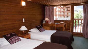 A wooden cabin room with two beds, a desk, and a balcony with a view of a building.