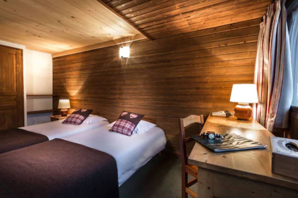 A wooden cabin room with two beds, a desk with a lamp, and a small window.