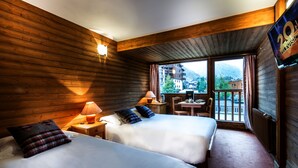 A hotel room with wooden walls, two beds, a TV, a balcony with a view of mountains, and a table with chairs. - Les Crêtes Blanches (Val-d'Isere)