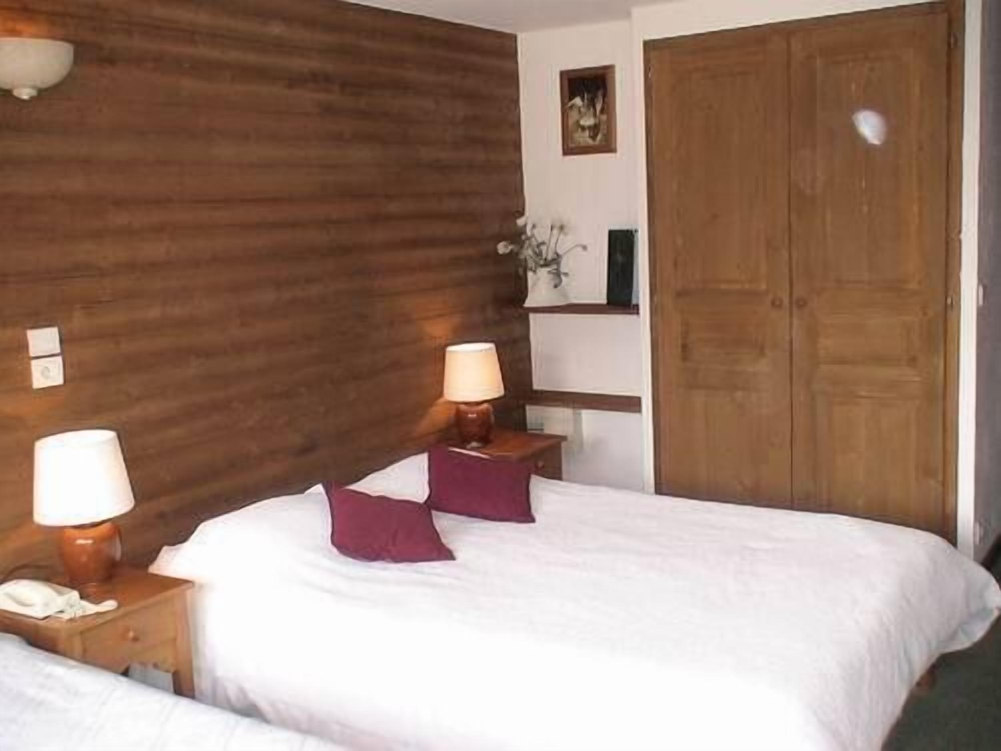 A bedroom with a wooden headboard, a bed with white bedding and two purple pillows, a nightstand with a lamp, and a wooden door.