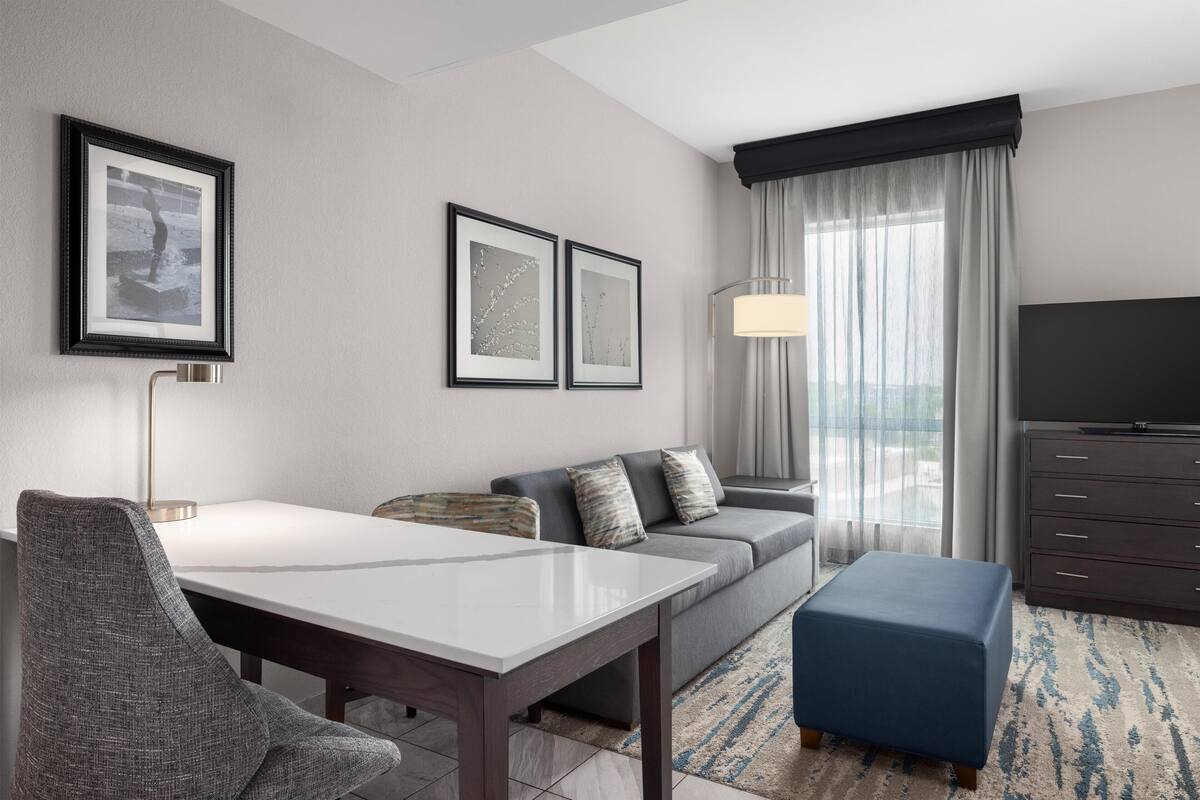 A modern hotel room with a dining table, sofa, ottoman, and a TV.