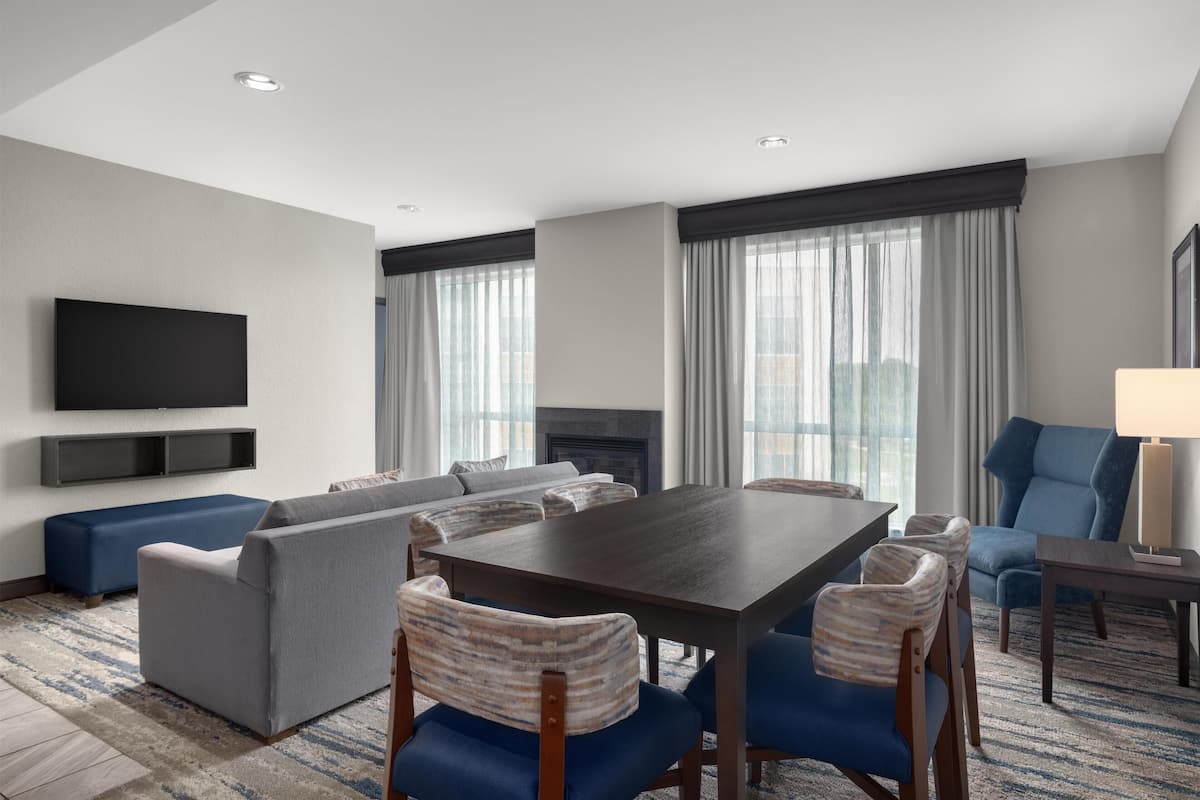A modern hotel room with a dining area, sofa, chairs, a flat-screen TV, and a fireplace.