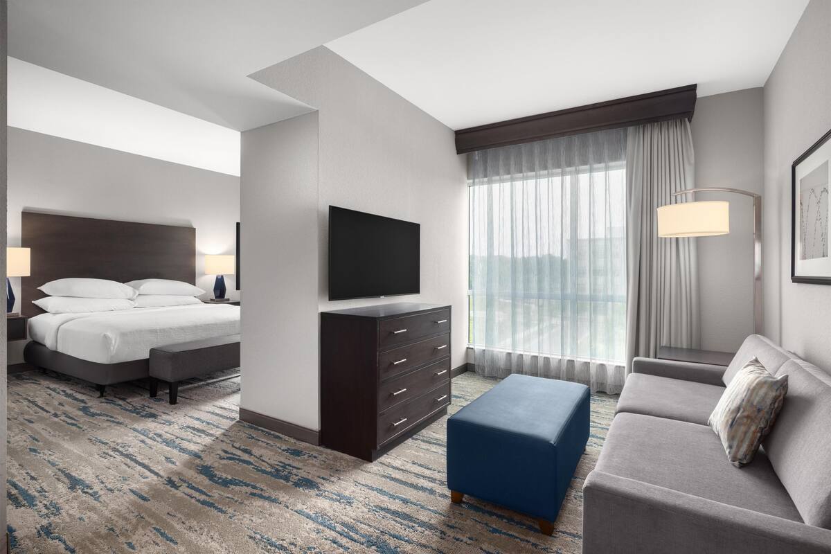 A hotel room with a bed, a TV, a dresser, a sofa, and a large window with curtains.