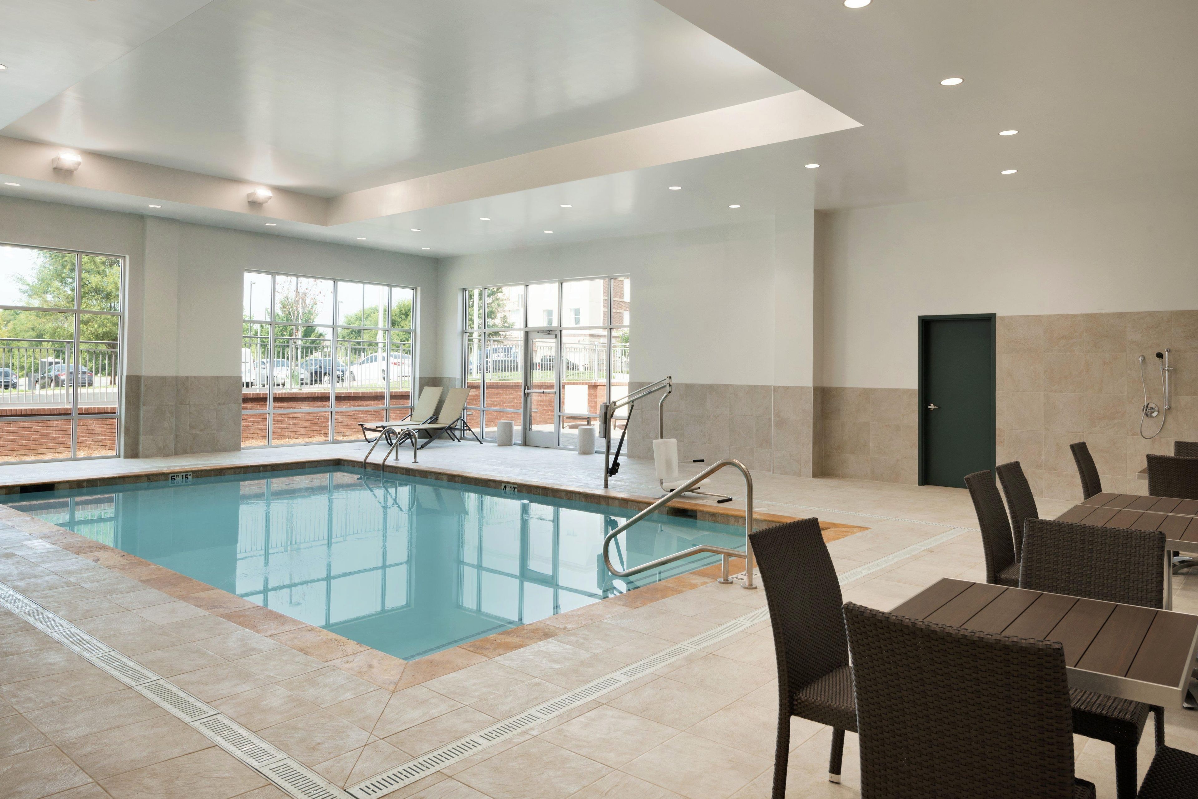 An indoor swimming pool with clear glass walls, a lounge area with chairs, and a dining area with tables and chairs.