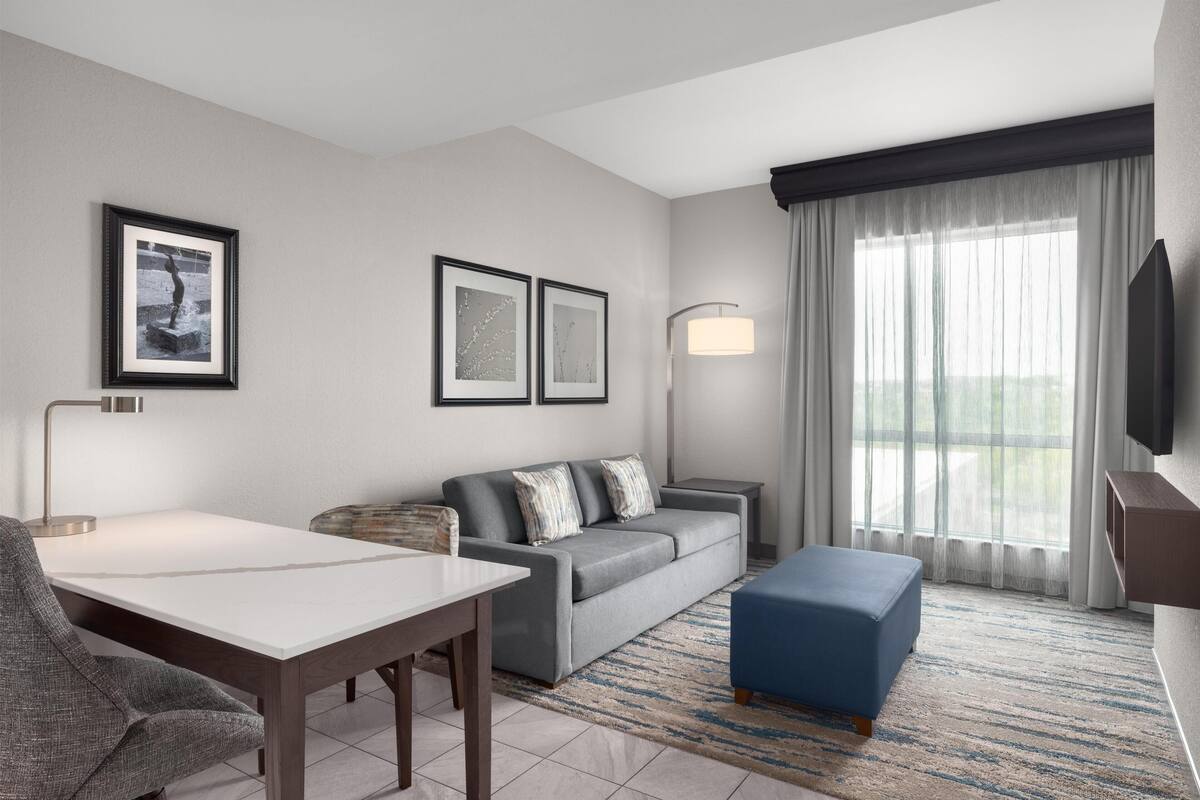 A modern hotel room with a sofa, a dining table, a blue ottoman, a TV, and framed artwork on the wall.