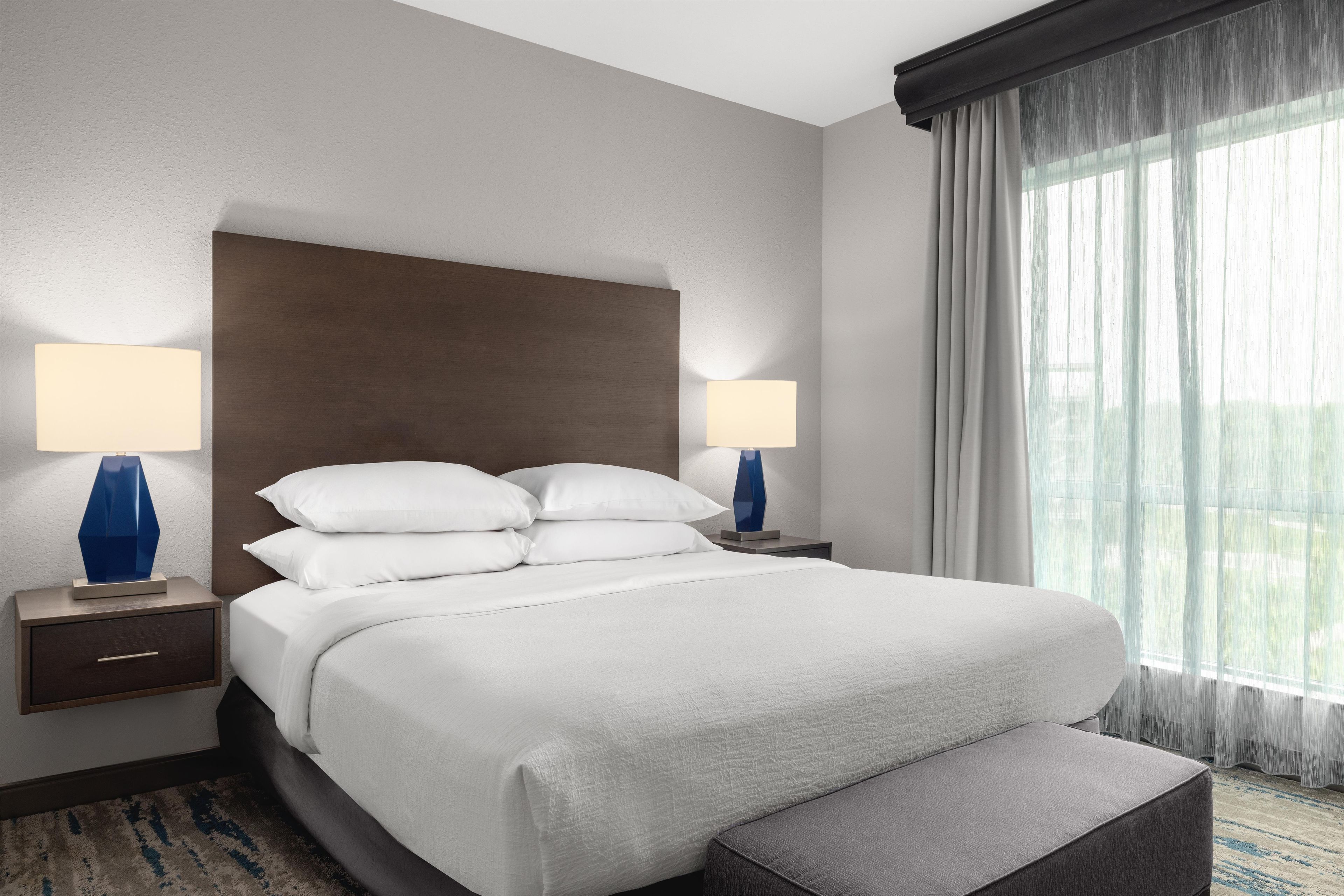 Habitación de hotel con una cama grande, dos mesitas de noche con lámparas, cabecera y vistas al exterior a través de cortinas traslúcidas.