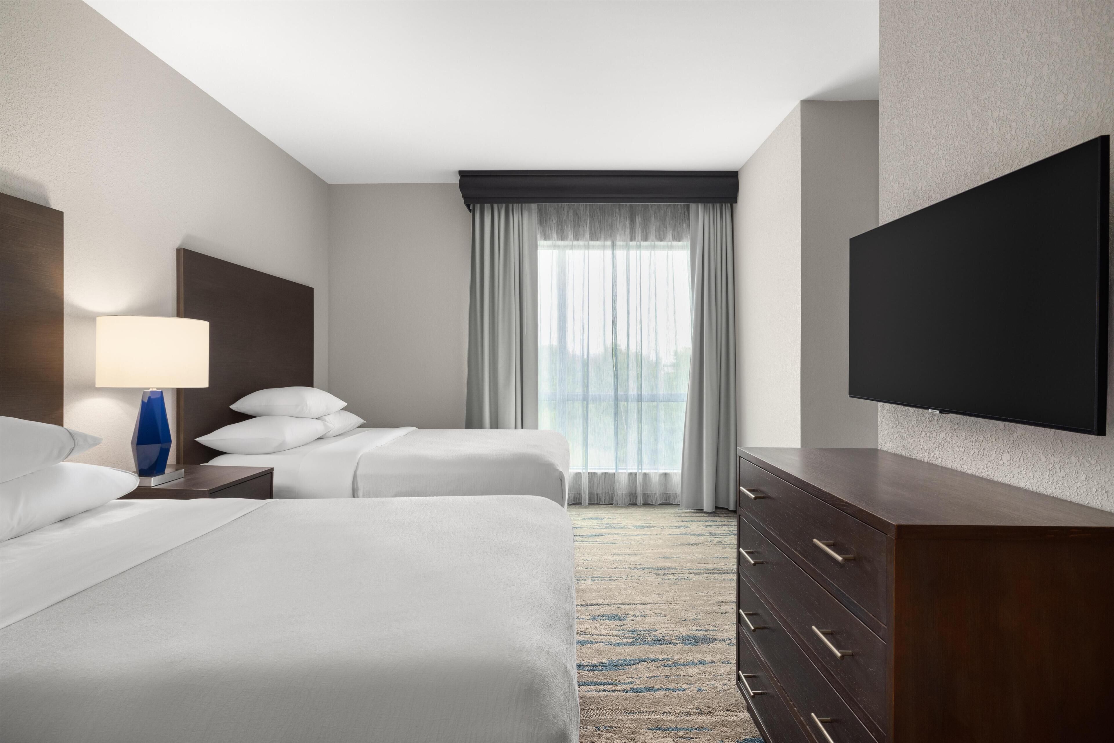 Une chambre d’hôtel avec deux lits, un téléviseur à écran plat, une table de chevet avec une lampe, une commode et une vue sur la ville à travers des rideaux transparents.