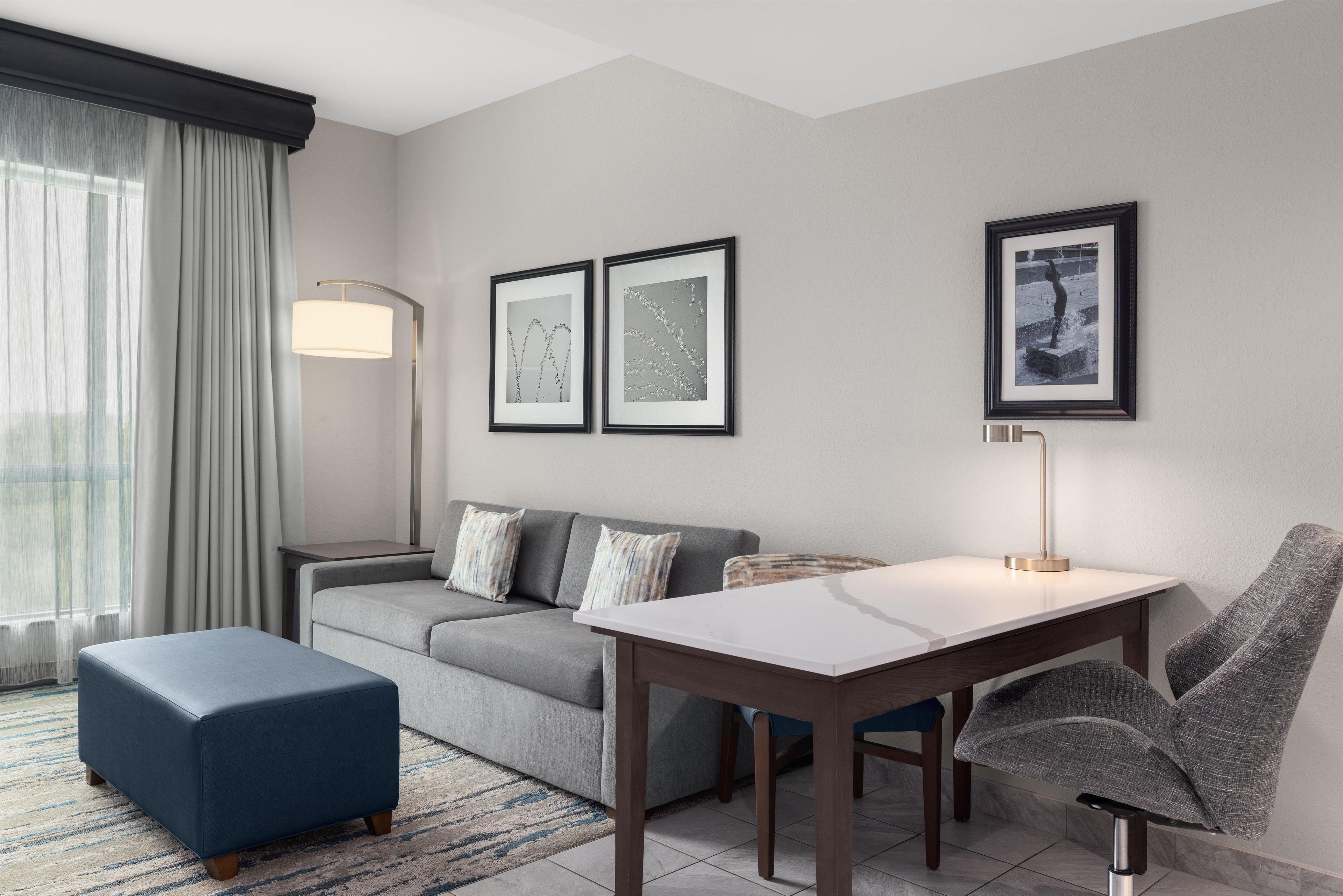 A modern hotel room with a grey sofa, a blue ottoman, a wooden dining table, and a grey armchair. There are two framed pictures on the wall, a floor lamp, and a window with curtains.
