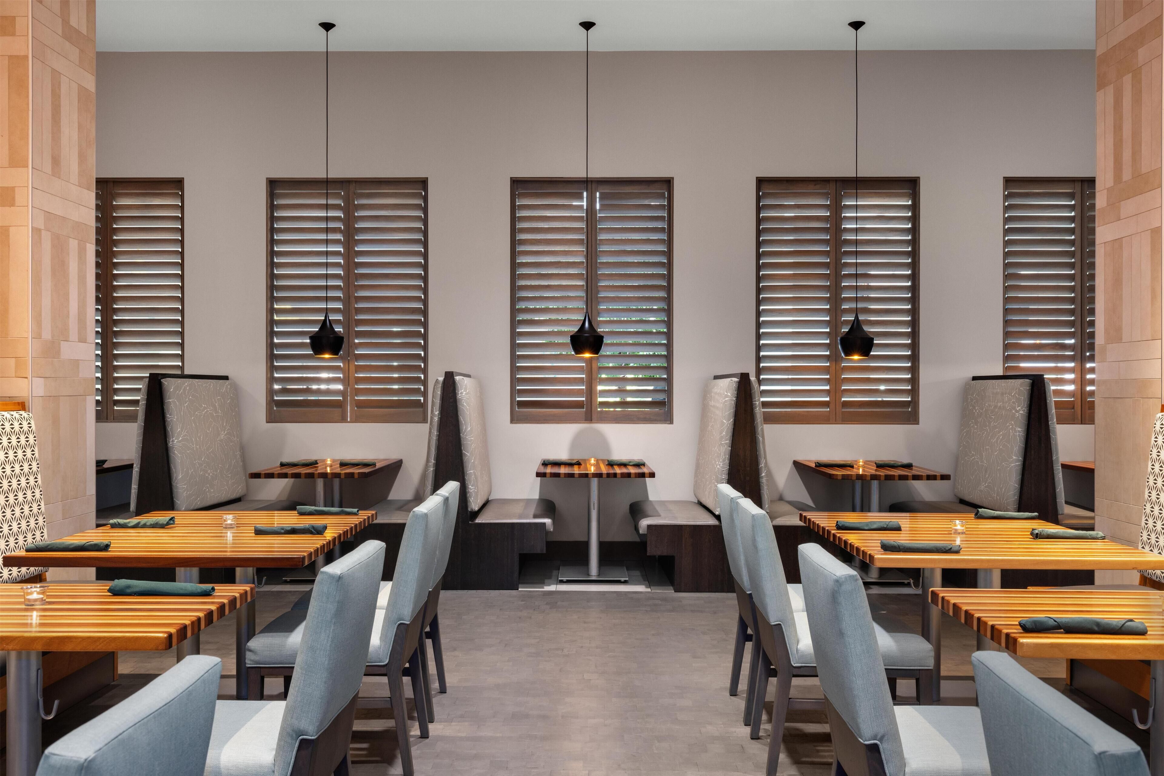 Un restaurante con mesas y sillas de madera, luces colgantes y grandes ventanales con persianas.