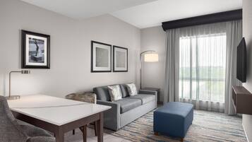 A modern hotel room with a grey sofa, a blue ottoman, a white table, and a flat-screen TV mounted on the wall.