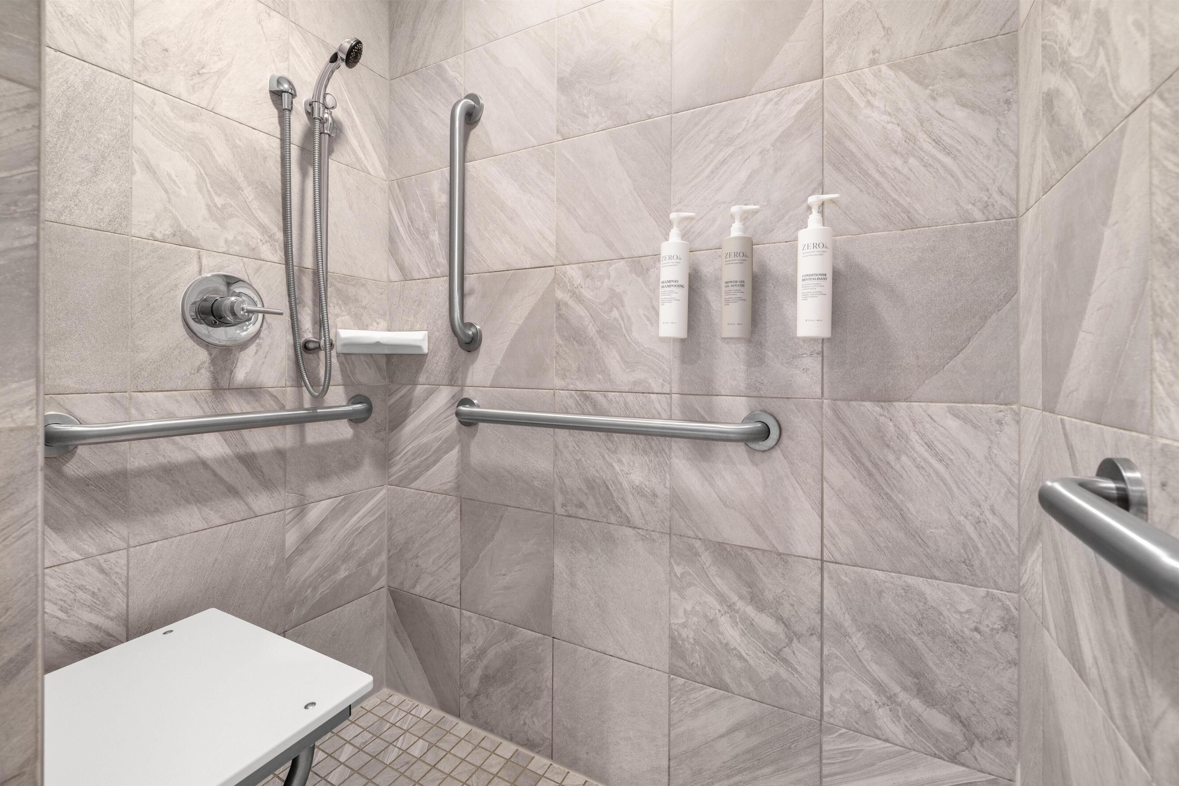 A bathroom with marble tiles, a shower with a handheld showerhead, grab bars, and two bottles of shampoo.