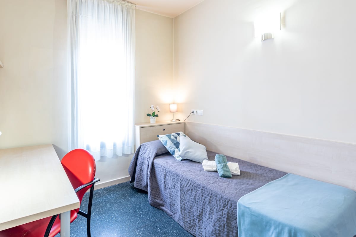 A small, neatly arranged hotel room with a single bed, a desk, and a chair.