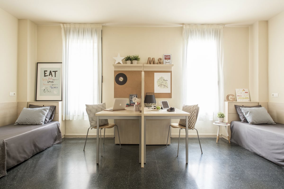 A room with a bed, a desk, and a chair. There is a framed picture on the wall and a small table with a plant.