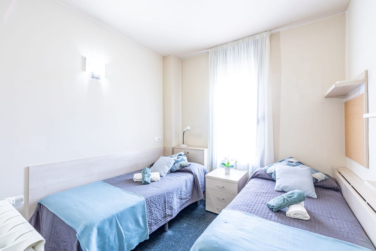 A twin room with two beds, a bedside table, a lamp, and a window with curtains.