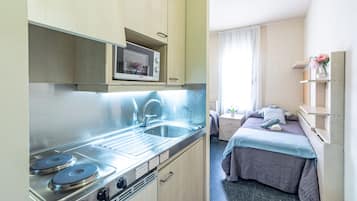 Una cucina compatta con forno a microonde, lavello e piano cottura. Si intravede un piccolo angolo ristoro con tavolo e sedie. La stanza presenta un letto con coperta blu e un comodino con un vaso di fiori.