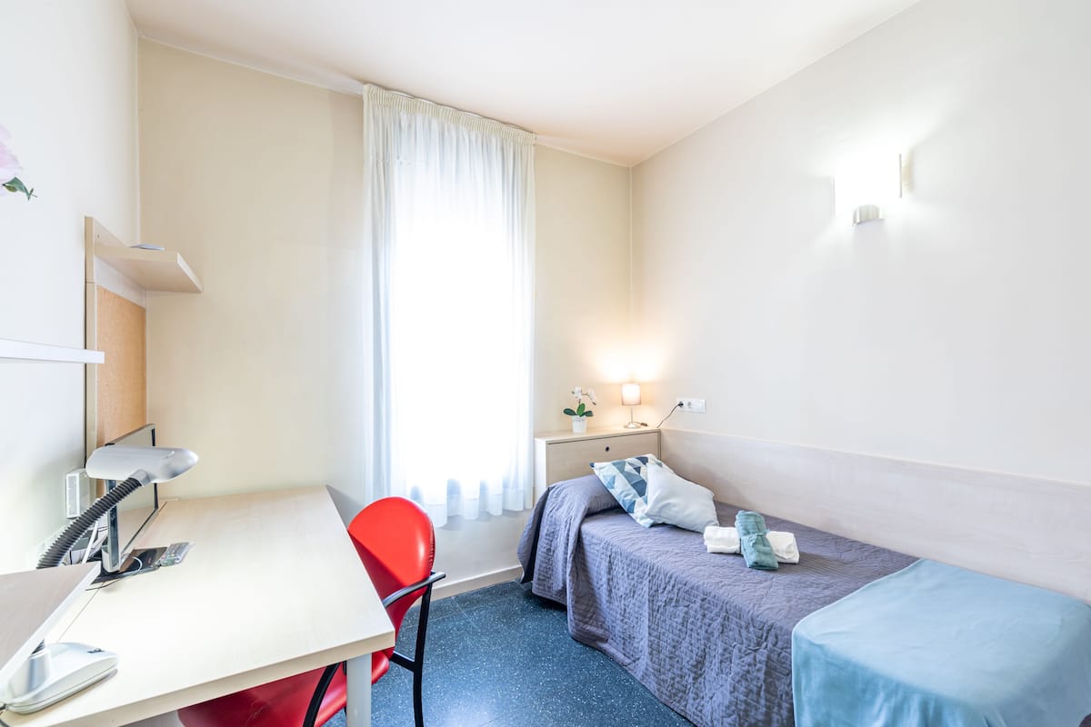 A small, well-lit room with a desk, a red chair, a bed with a blue bedspread, and a nightstand with a lamp.