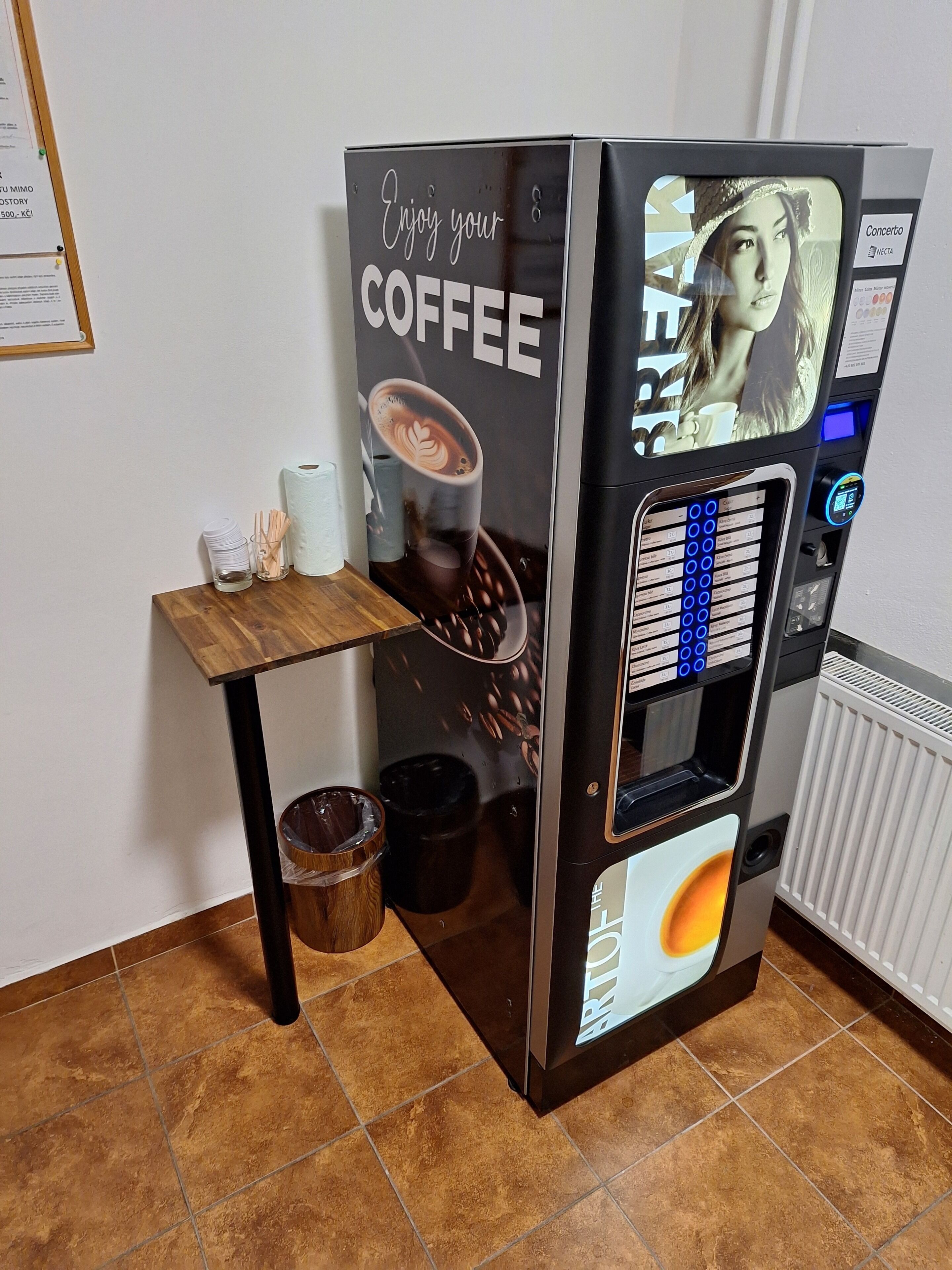 Un distributeur de café orné d’un motif représentant une tasse de café, une petite table avec des tasses et des serviettes en papier, et une poubelle.