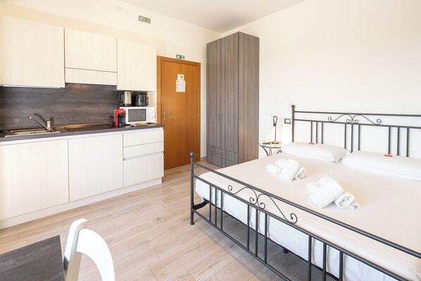 Een hotelkamer met een bed, een klein tafeltje met een lamp, een kitchenette met een magnetron, en een houten garderobe.