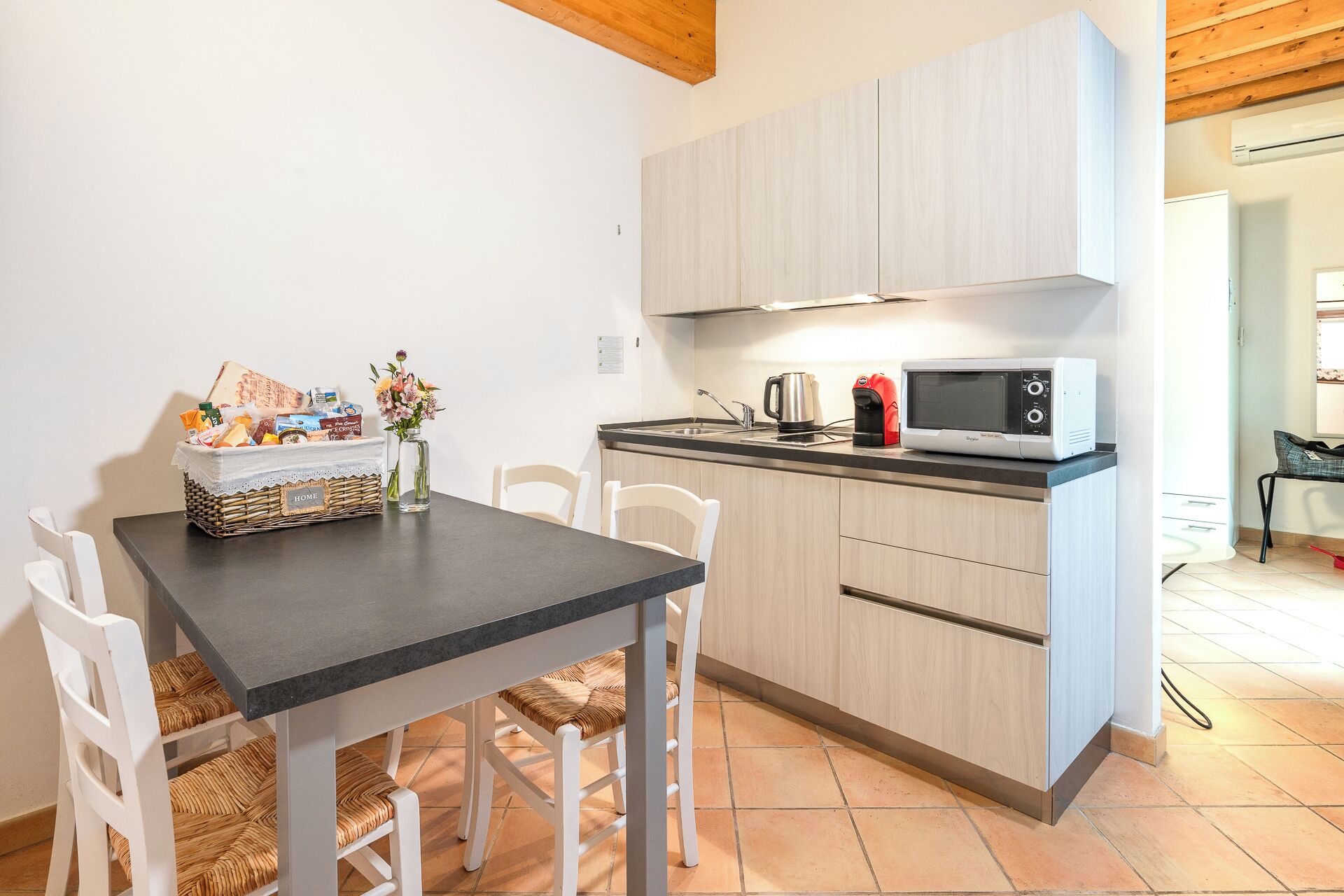 A compact kitchen with a dark countertop, white cabinetry, and a microwave. There is a wicker basket with food items and a vase with flowers on the table.