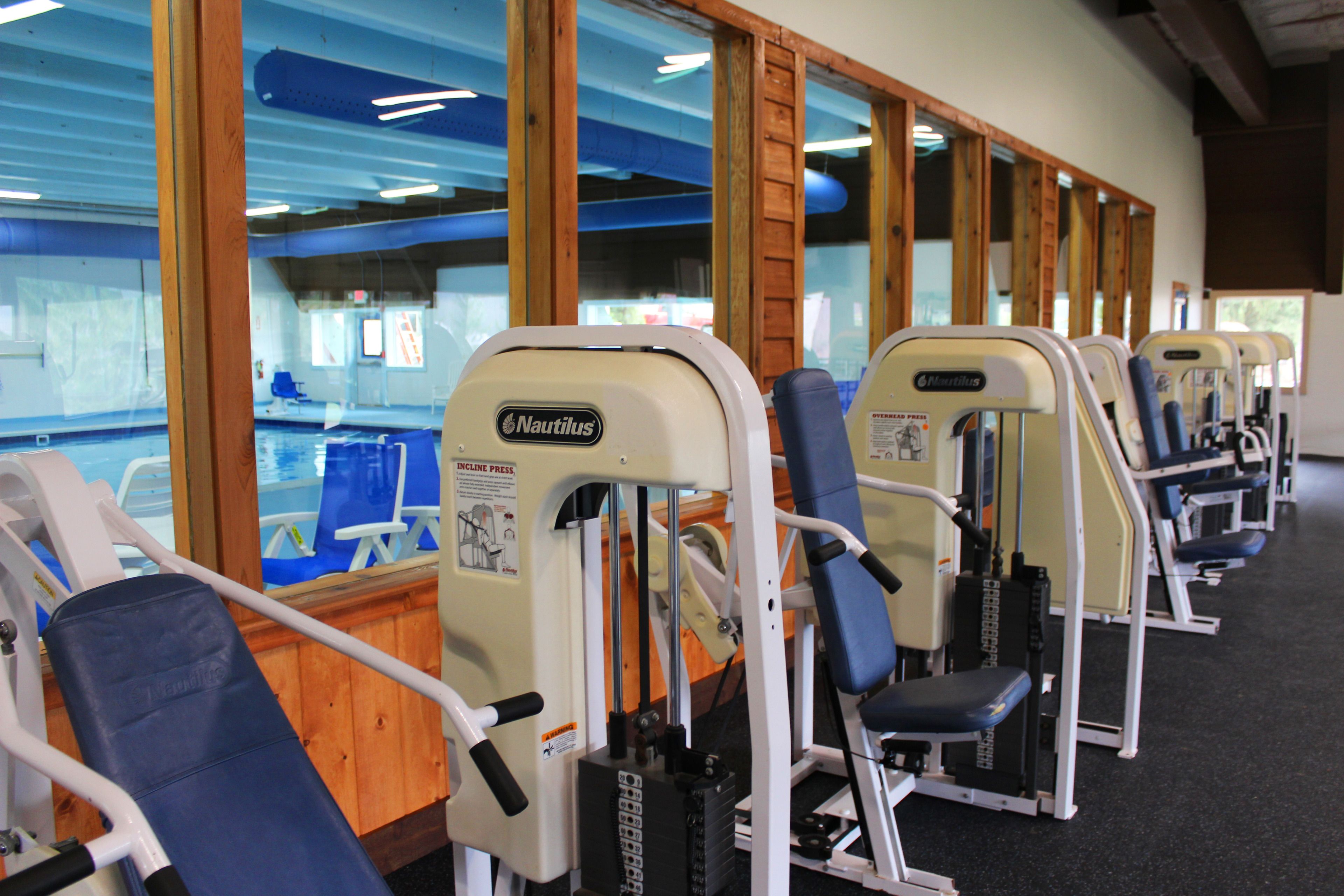 Varias máquinas de ejercicio Nautilus en un gimnasio con amplios ventanales que dan a una zona de piscina.