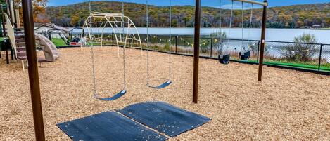 A playground with swings and a slide, surrounded by a fence and a lake view.