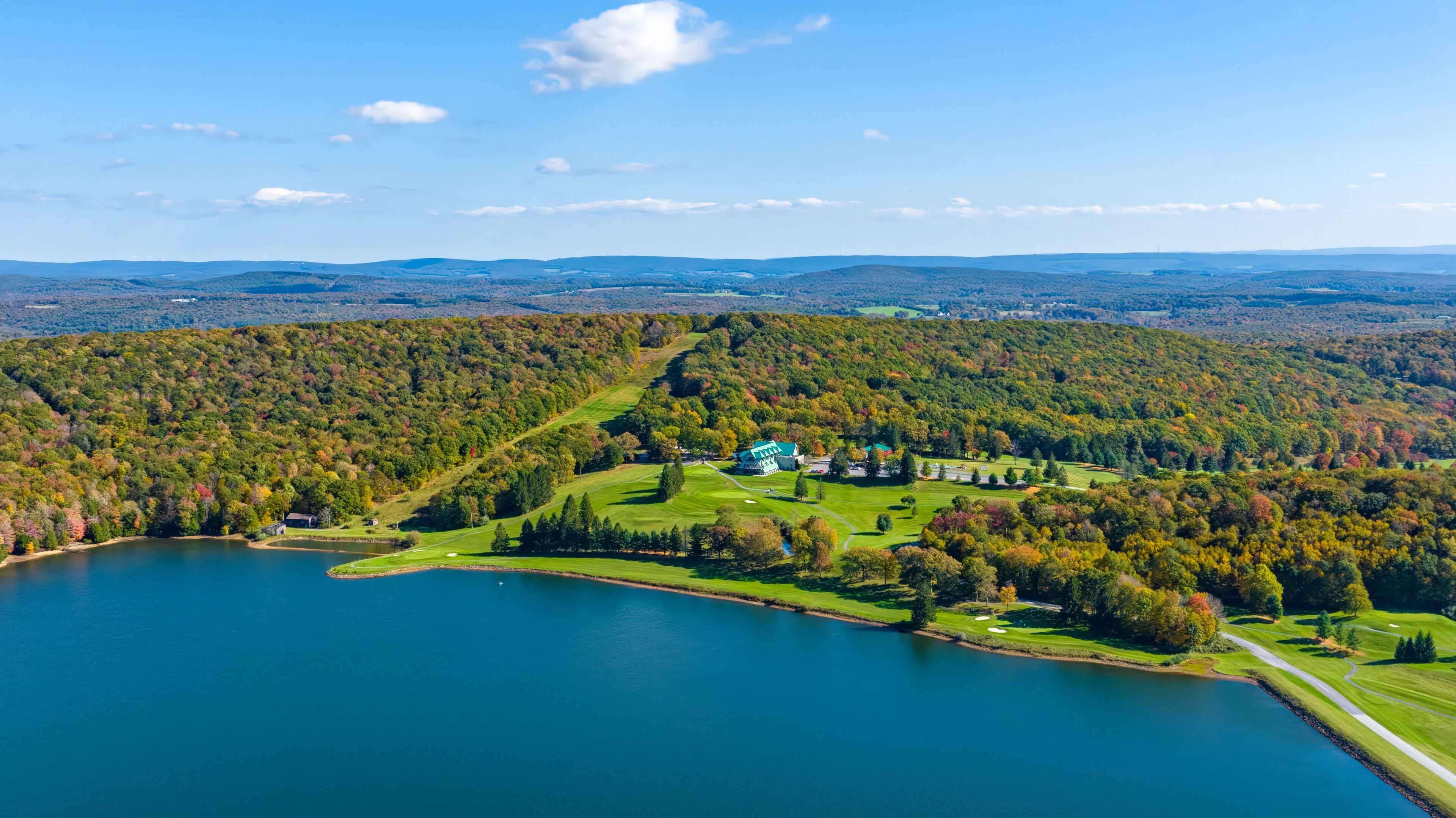 Un lago extenso rodeado de colinas boscosas, un campo de golf y un edificio con techo verde.