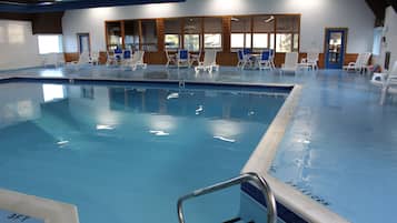 Una piscina interior con agua azul cristalina, rodeada de sillas reclinables blancas y una terraza de madera.