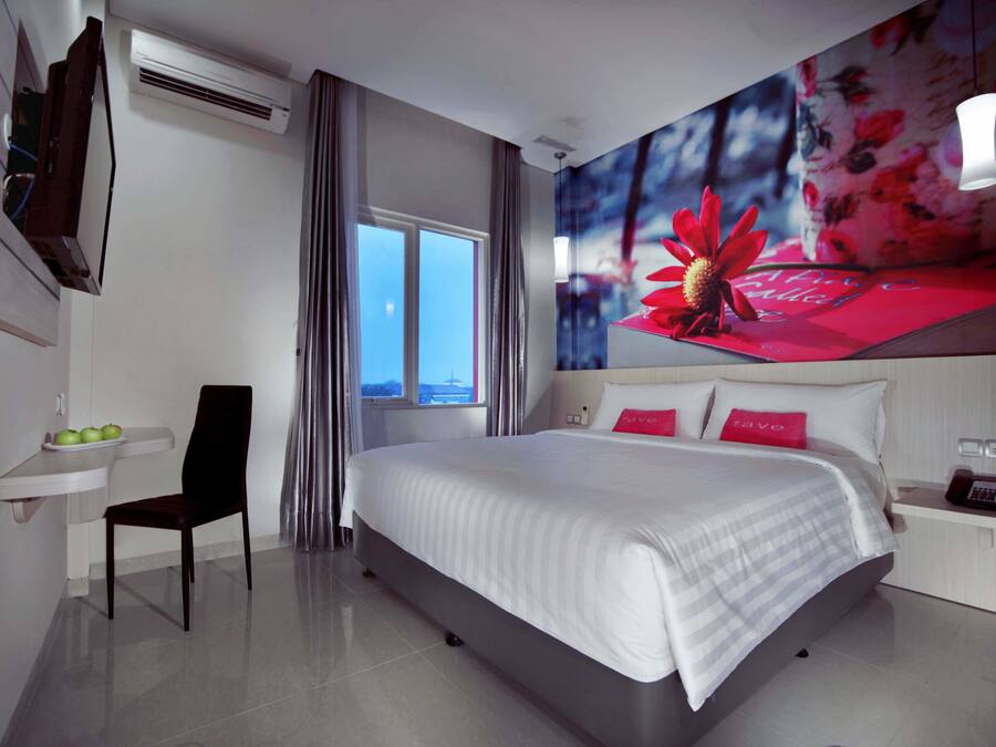 A modern hotel room with a large bed, a desk with a chair, a small table with apples, and a window with curtains.