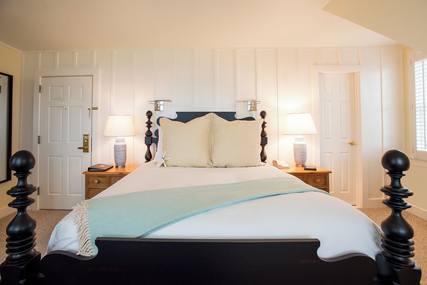Una cama bien hecha con una colcha blanca y azul claro, flanqueada por dos mesitas de noche con lámparas, en una habitación con paredes blancas de paneles y un cabecero de madera oscura.