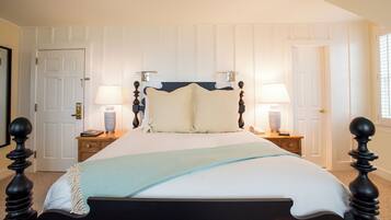 Una cama bien hecha con una colcha blanca y azul claro, flanqueada por dos mesitas de noche con lámparas, en una habitación con paredes blancas de paneles y un cabecero de madera oscura.