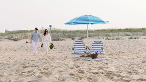 A sandy beach with two people walking, a blue and white striped beach umbrella, and two striped beach chairs. - The Wauwinet (Nantucket)