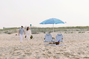 Una playa arenosa con dos personas caminando, una sombrilla de playa azul y blanca a rayas, y dos sillas de playa a rayas.