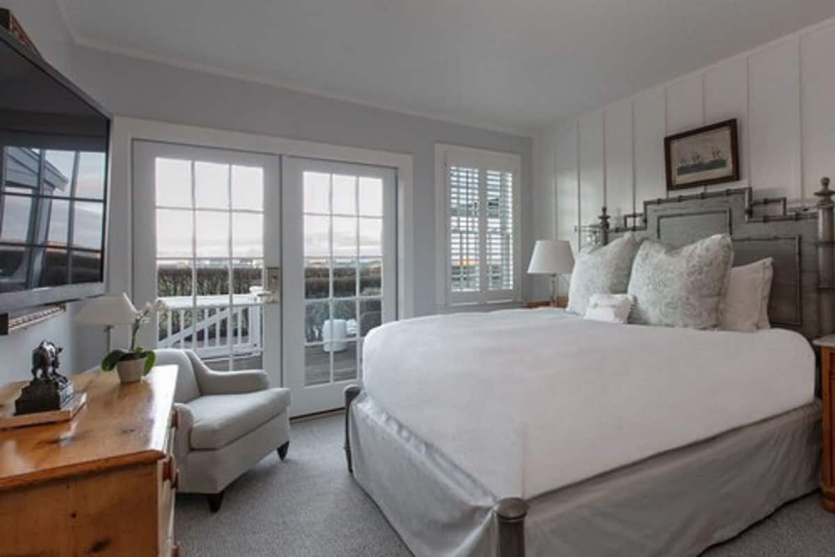 Une chambre avec un grand lit, un bureau, une chaise et un téléviseur. Elle est dotée de fenêtres et d’un balcon.