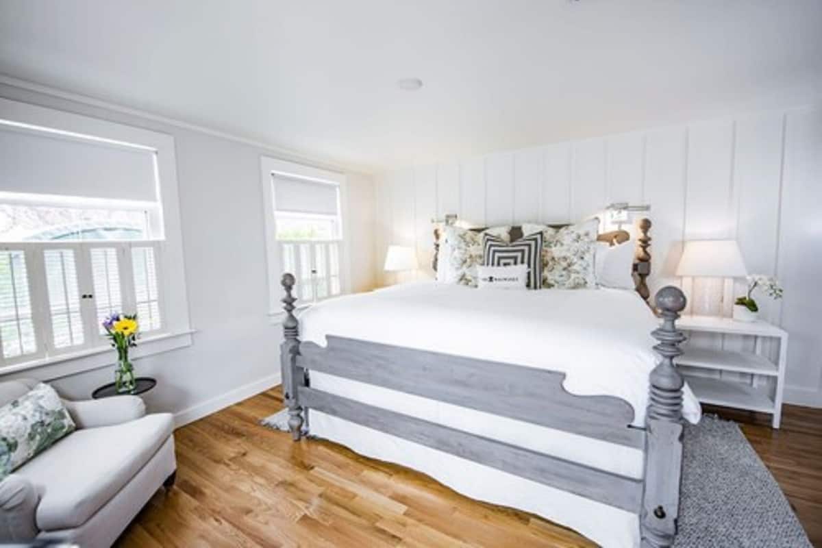 Une chambre à coucher avec un grand lit, une table de chevet, une lampe, une fenêtre et une petite table ornée de fleurs.