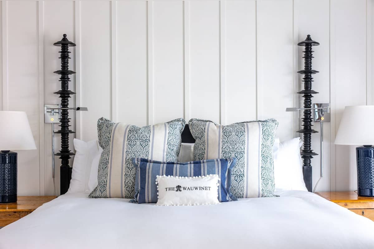 Un lit bien fait, agrémenté d’oreillers à motifs et d’un coussin décoratif avec un texte, flanqué de deux lampes hautes, noires et de style spiralé, placées de chaque côté.