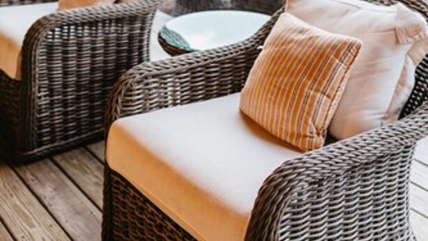 A covered outdoor seating area with wicker furniture and cushions, large windows, and a view of greenery. - The Wauwinet (Nantucket)
