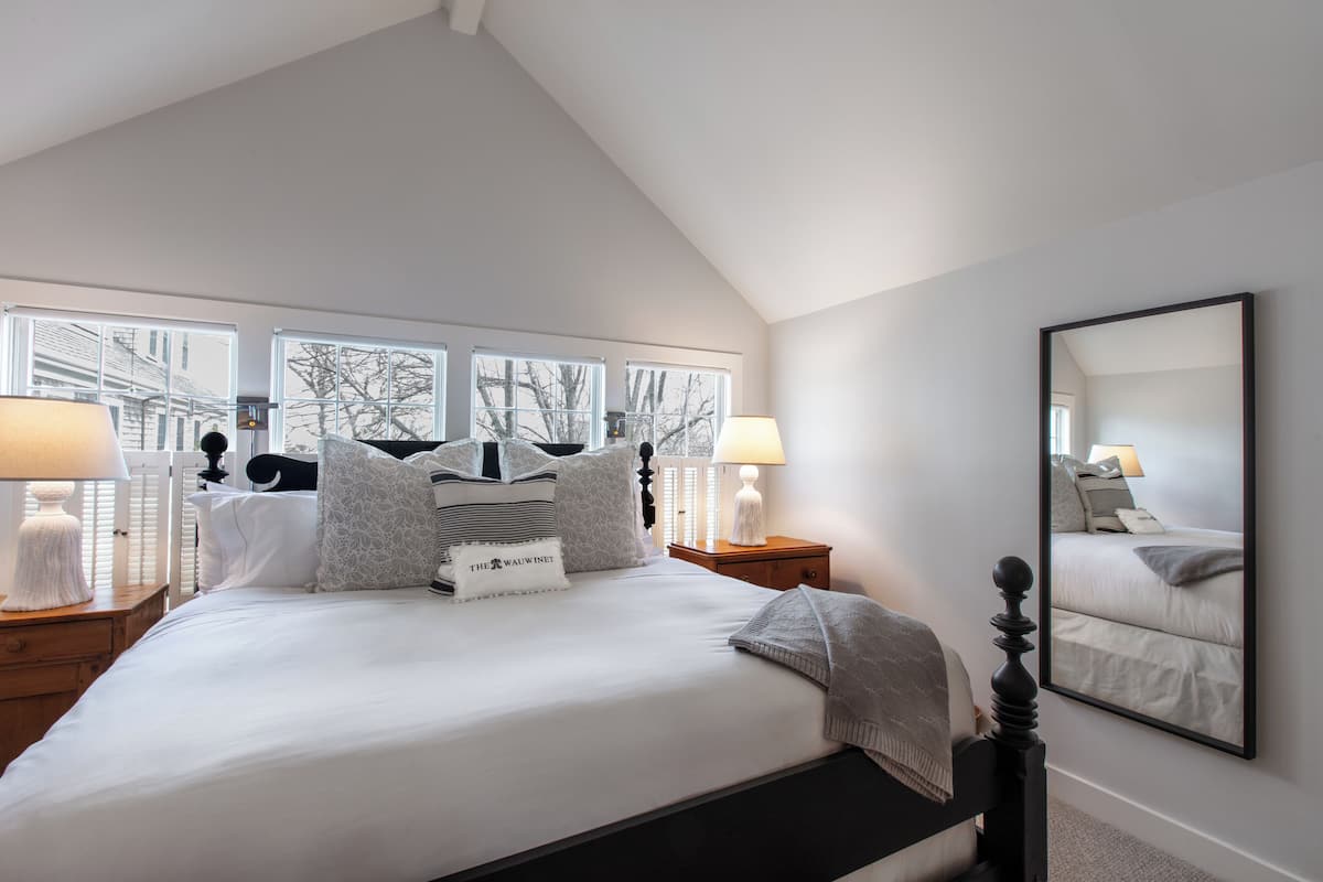 Une chambre avec un grand lit, deux tables de chevet, un miroir et une vue sur les arbres par les fenêtres.