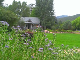 A house with a grey roof and a porch, surrounded by a green lawn and purple flowers.