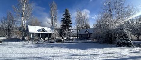 A snow-covered landscape with two houses, one with a gabled roof and another with a similar roof, surrounded by bare trees and a clear sky.