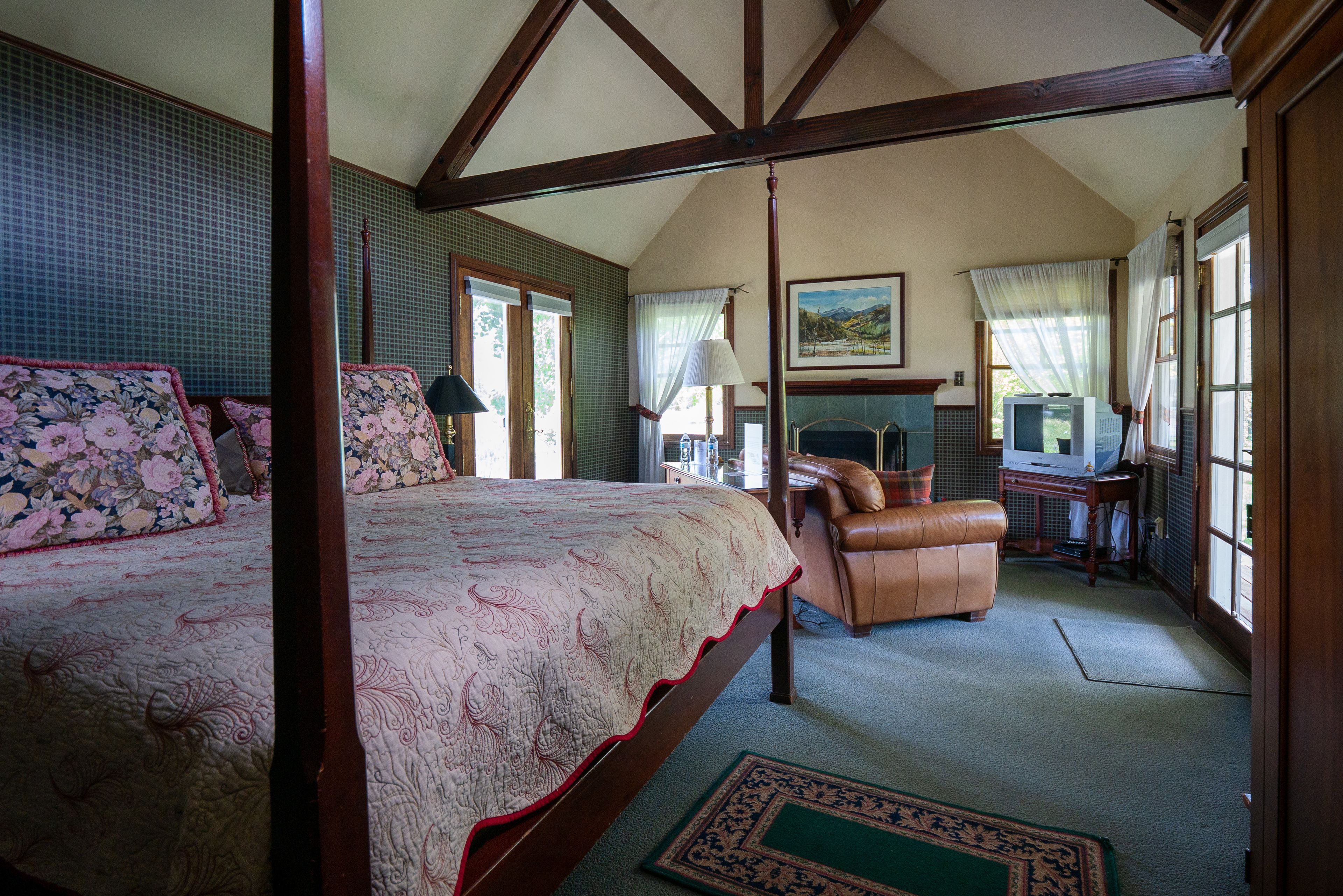 A bedroom with a four-poster bed, a desk with a chair, and a view of the outdoors through large windows.