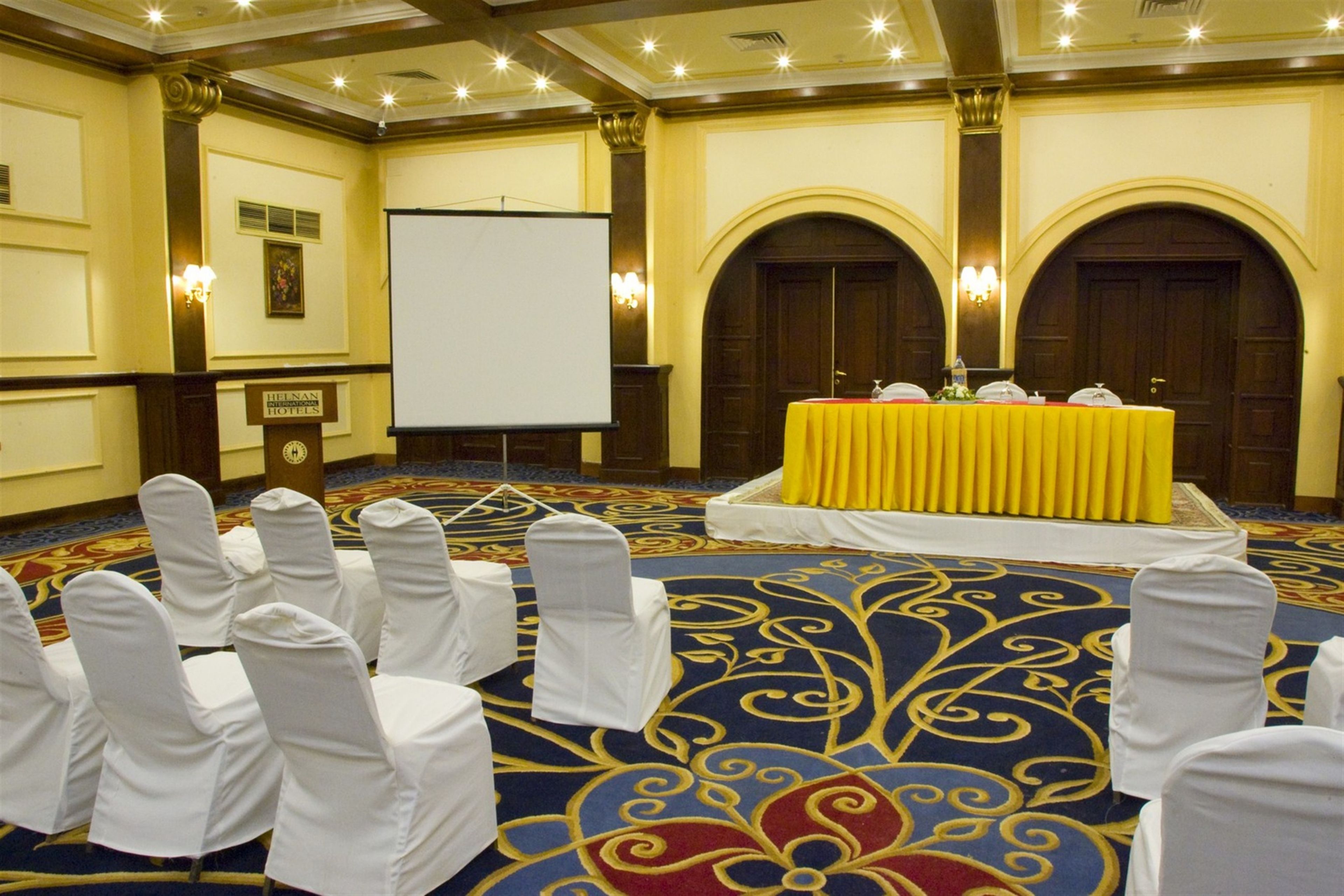 A conference room with a stage, white chairs, a carpeted floor, and a projector screen.