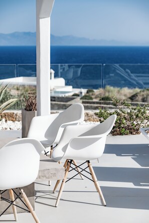 A modern outdoor seating area with white chairs and a table, overlooking a scenic view of the sea and mountains. - Villa Alice (Mykonos)
